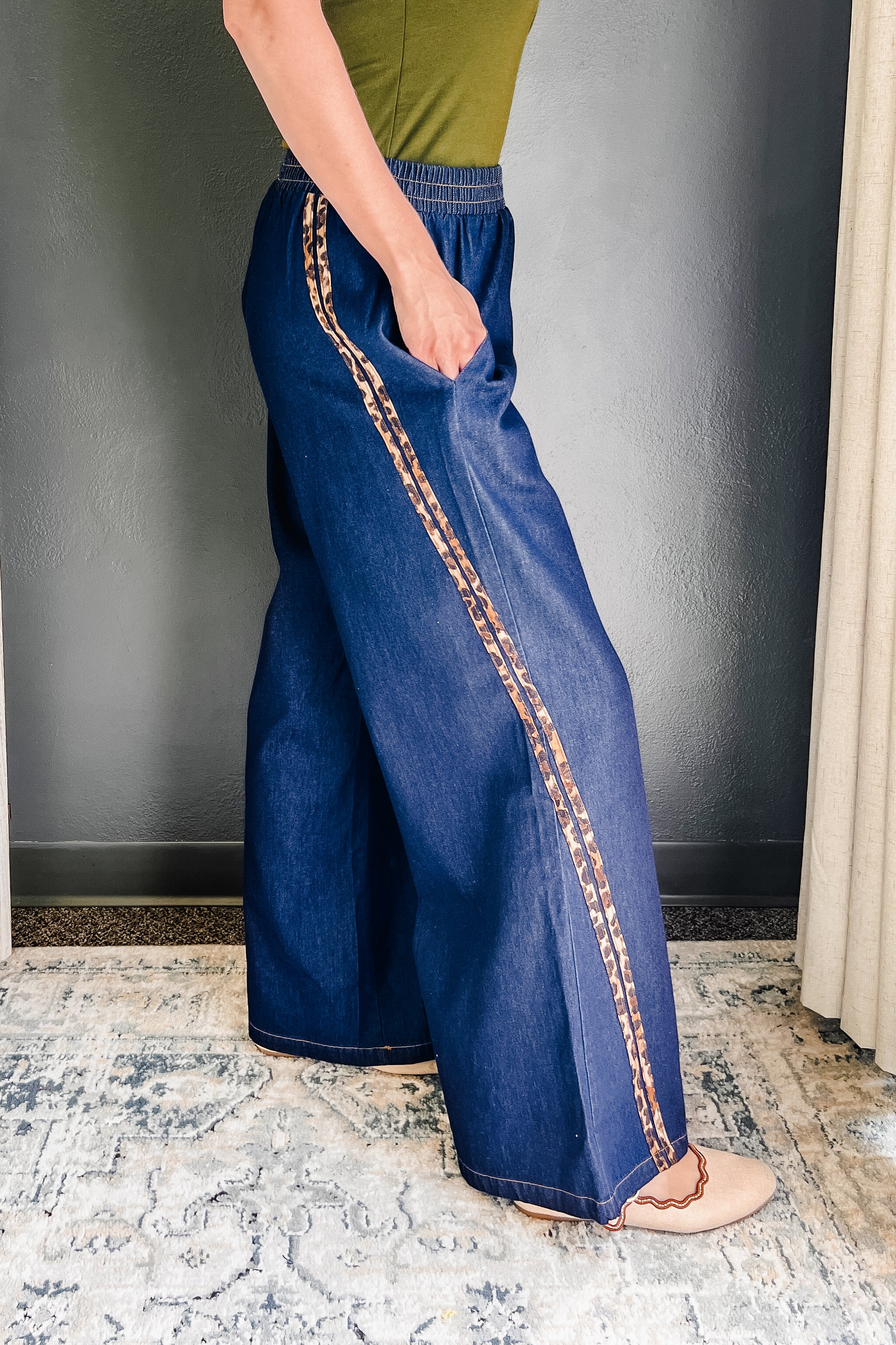 A person wearing wide-leg blue denim pants with  cheetah stripe running down the side, paired with a green top and beige sandals, standing on a patterned rug.