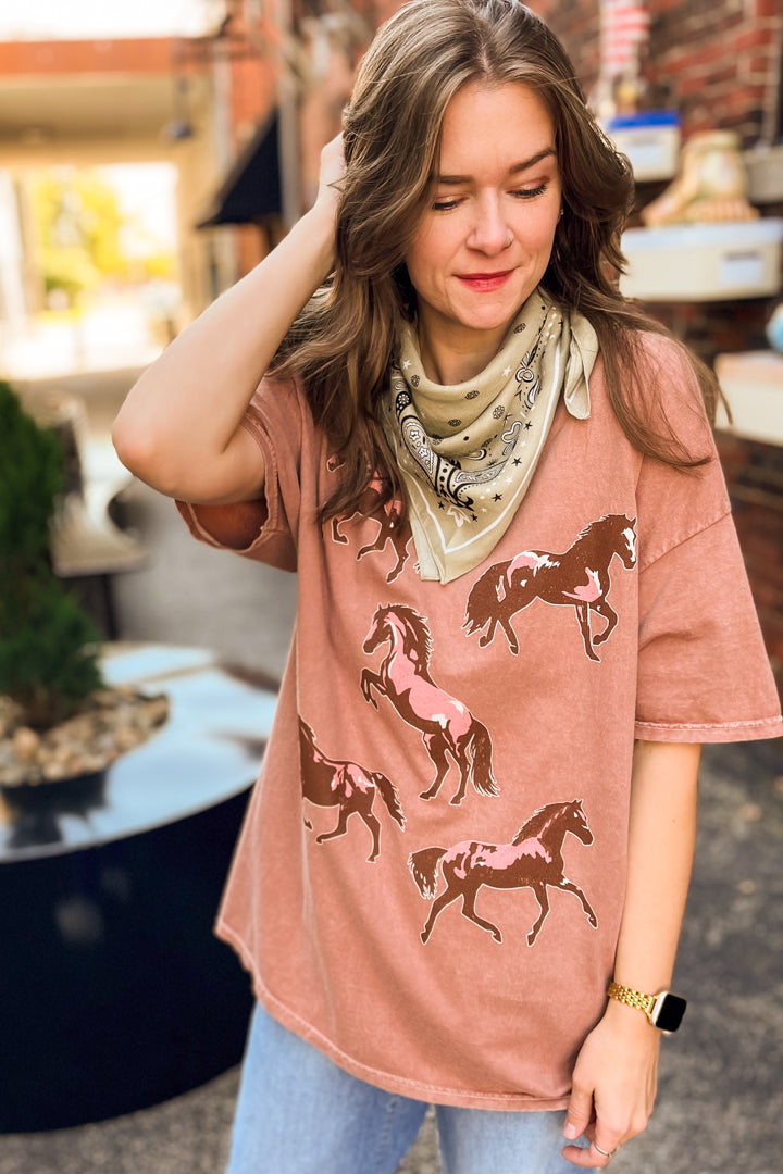 A woman wearing a dusty rose short-sleeve t-shirt with a brown and white horse print, styled with a beige bandana and blue jeans in an outdoor setting.