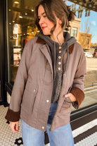 A woman wearing a brown and beige houndstooth jacket with brown ribbed trim over a gray button-front top and light blue jeans, standing in front of a storefront window.