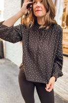 A woman wearing a black long-sleeve blouse with small white polka dots and a tie-neck detail, paired with black leggings, standing outdoors.