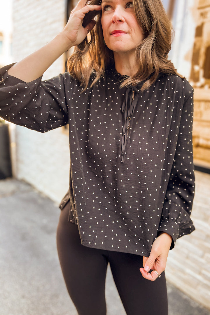 A woman wearing a black long-sleeve blouse with small white polka dots and a tie-neck detail, paired with black leggings, standing outdoors.