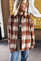 A woman wearing a rust-orange and beige plaid button-up flannel shirt over a brown graphic tee and blue jeans, standing in front of a yellow wall with a white boot graphic.
