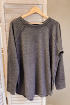 A dark gray heathered knit long-sleeve top with raglan sleeves hanging on a wooden hanger against a light wood slatted backdrop.