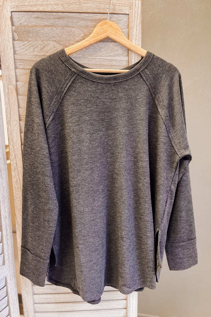 A dark gray heathered knit long-sleeve top with raglan sleeves hanging on a wooden hanger against a light wood slatted backdrop.