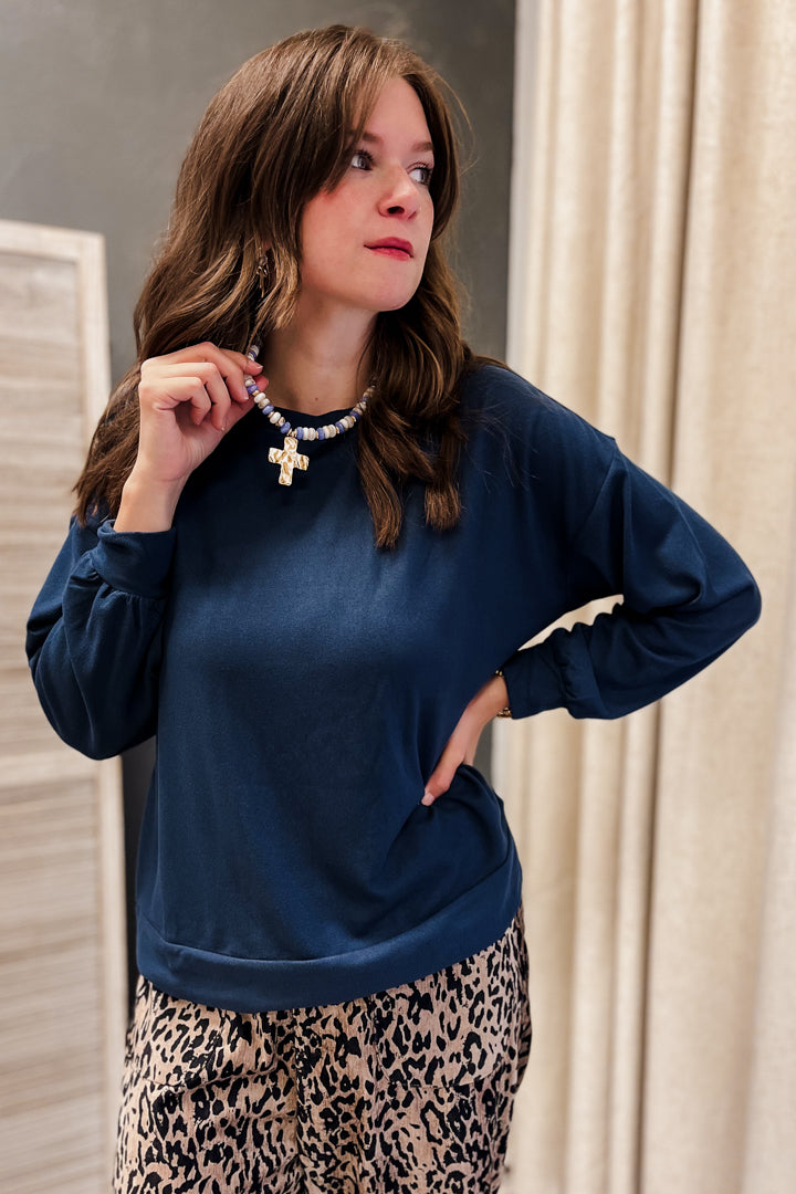 A woman wearing a navy blue long-sleeve top and a leopard print skirt, accessorized with a beaded necklace featuring a gold cross pendant, posing in front of a neutral indoor background.