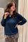 A woman wearing a navy blue long-sleeve top and a leopard print skirt, accessorized with a beaded necklace featuring a gold cross pendant, posing in front of a neutral indoor background.