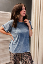 A woman wearing a light blue acid-wash short-sleeve t-shirt and leopard print pants, standing in an indoor setting with a gray wall and beige curtains.