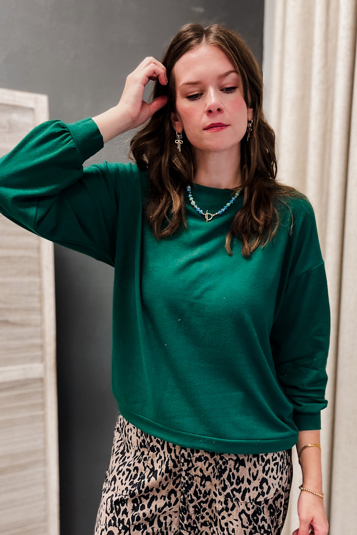 A woman wearing a dark green long-sleeve crewneck sweatshirt and a leopard print skirt, standing in front of a neutral indoor background.