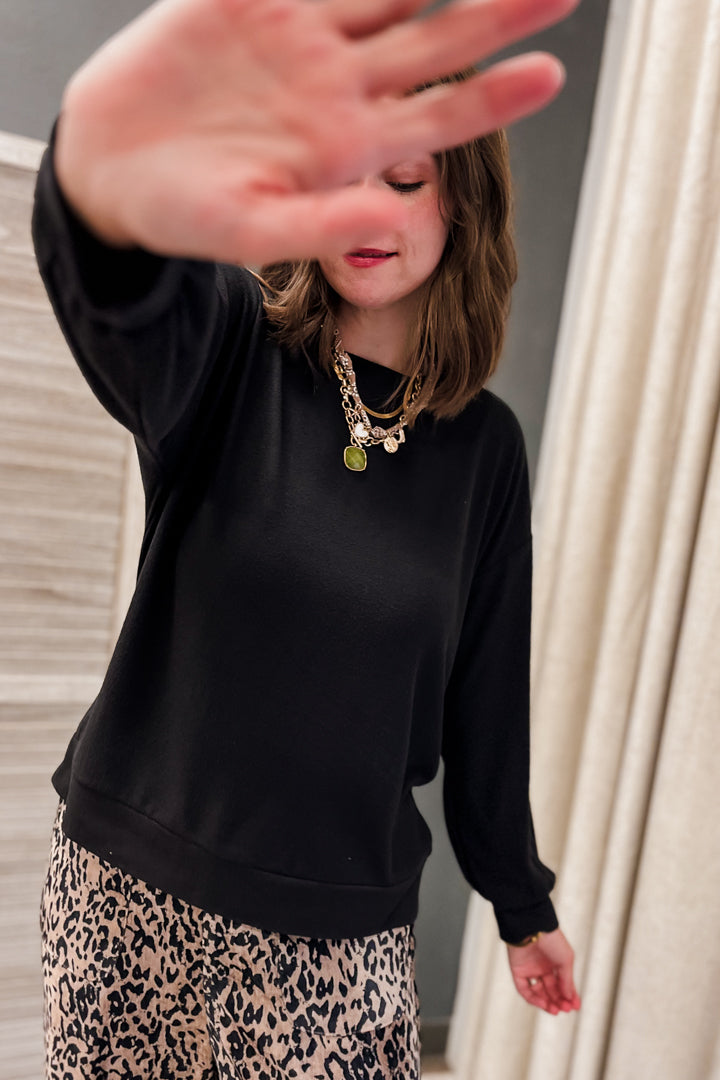 A woman wearing a black long-sleeve crewneck sweatshirt and a beige leopard print skirt, with layered gold necklaces, posing indoors with her hand partially covering the camera.