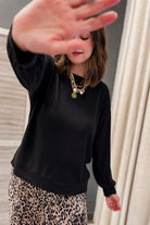 A woman wearing a black long-sleeve crewneck sweatshirt and a beige leopard print skirt, with layered gold necklaces, posing indoors with her hand partially covering the camera.