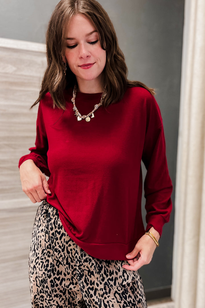 A woman wearing a long-sleeve crewneck sweatshirt in deep red, paired with a beige and black leopard print skirt, standing indoors.