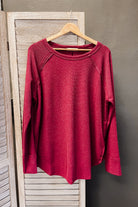 A heathered red long-sleeve raglan top hanging on a wooden hanger against a rustic light wood screen and dark gray wall.
