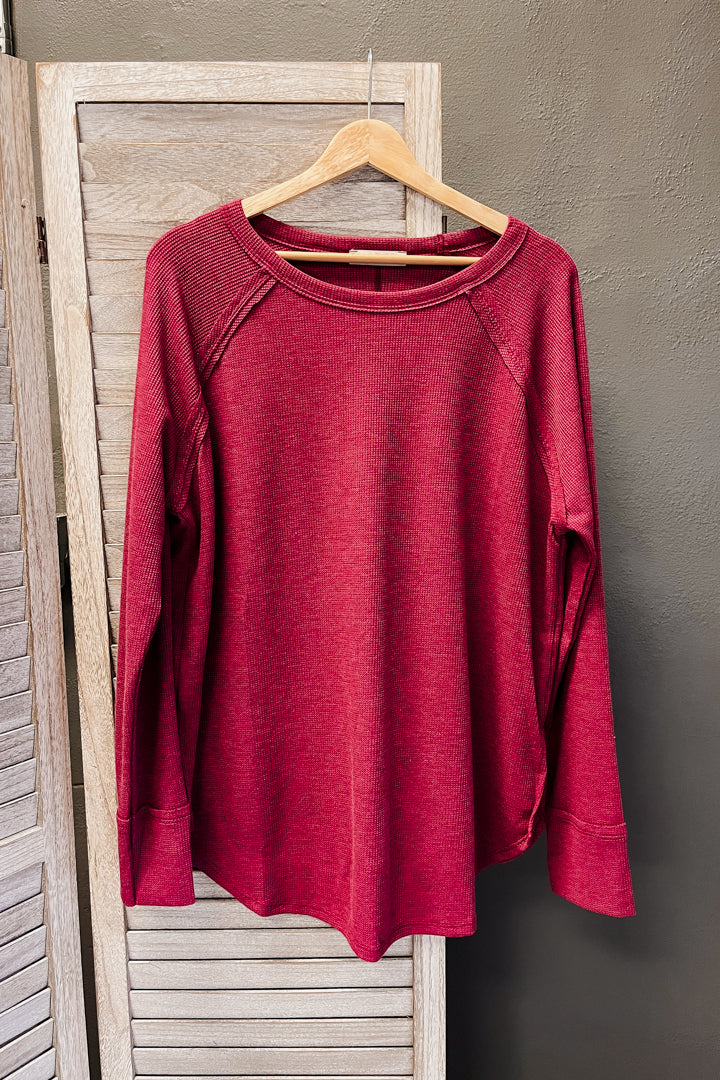 A heathered red long-sleeve raglan top hanging on a wooden hanger against a rustic light wood screen and dark gray wall.