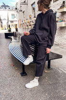 A person wearing a dark charcoal gray matching lounge set consisting of a pullover hoodie and wide-leg sweatpants, sitting on a curved black bench in an outdoor urban setting.