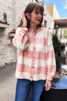 A woman wearing a pink and cream striped knit cardigan with blue jeans, standing outdoors in front of a stone building.