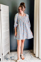 A woman wearing a light blue smocked dress with a pink and orange floral pattern, standing in a room with a dark gray wall, white shutter screen, and beige curtain.