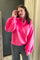 A woman wearing a bright neon pink long-sleeve half-placket pullover sweatshirt with buttoned cuffs, paired with dark gray pants, standing indoors against a dark gray wall and beige curtain background.