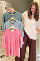 A woman stands indoors holding three ribbed knit short-sleeve tops on wooden hangers, shown in a lifestyle setting. The tops are in three different colors: dusty blue, light blue, and pink, layered on a neutral background.