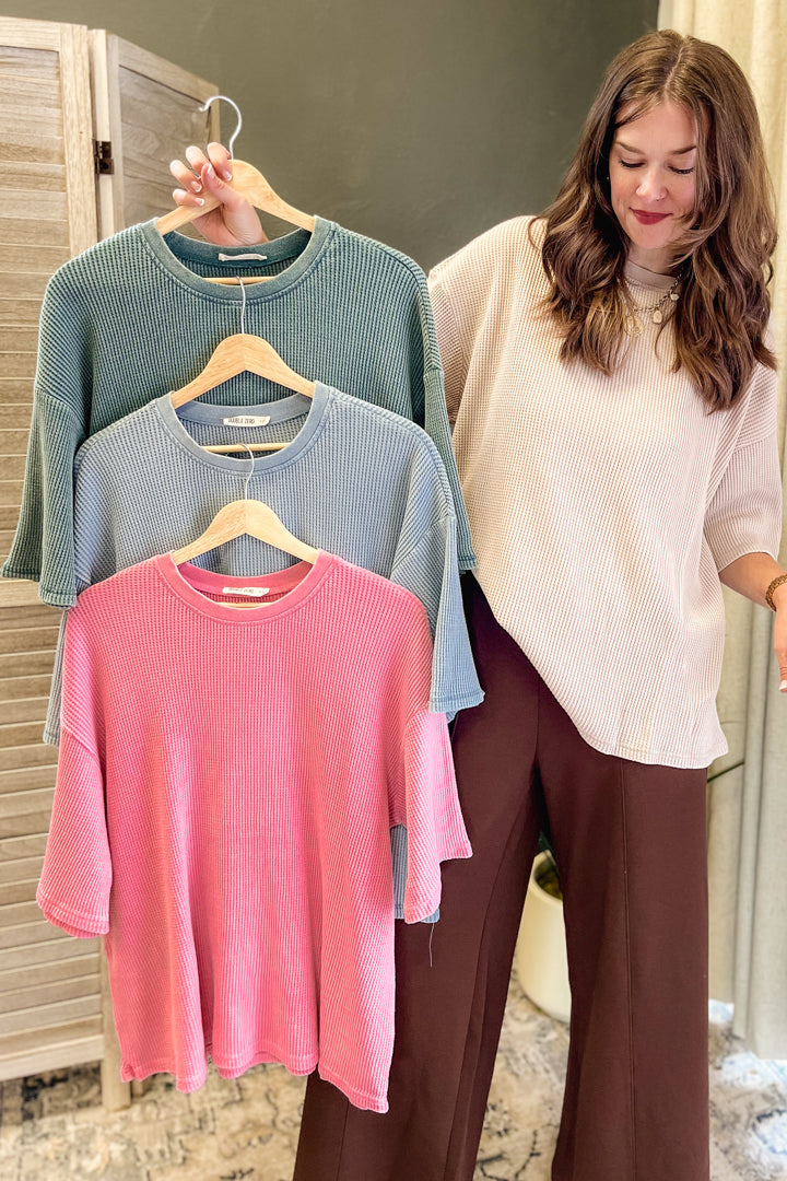 A woman stands indoors holding three ribbed knit short-sleeve tops on wooden hangers, shown in a lifestyle setting. The tops are in three different colors: dusty blue, light blue, and pink, layered on a neutral background.