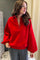 A woman wearing a bright red long-sleeve henley-style top with a buttoned collar and gray pants, standing indoors against a neutral curtain and wall background.