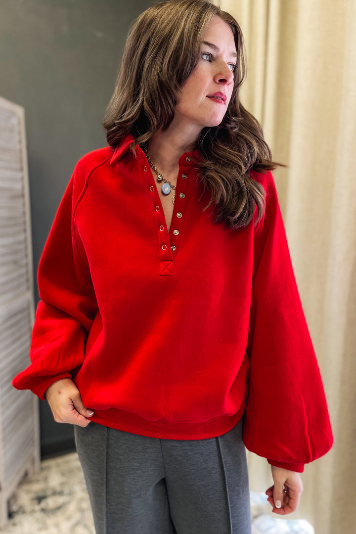 A woman wearing a bright red long-sleeve henley-style top with a buttoned collar and gray pants, standing indoors against a neutral curtain and wall background.
