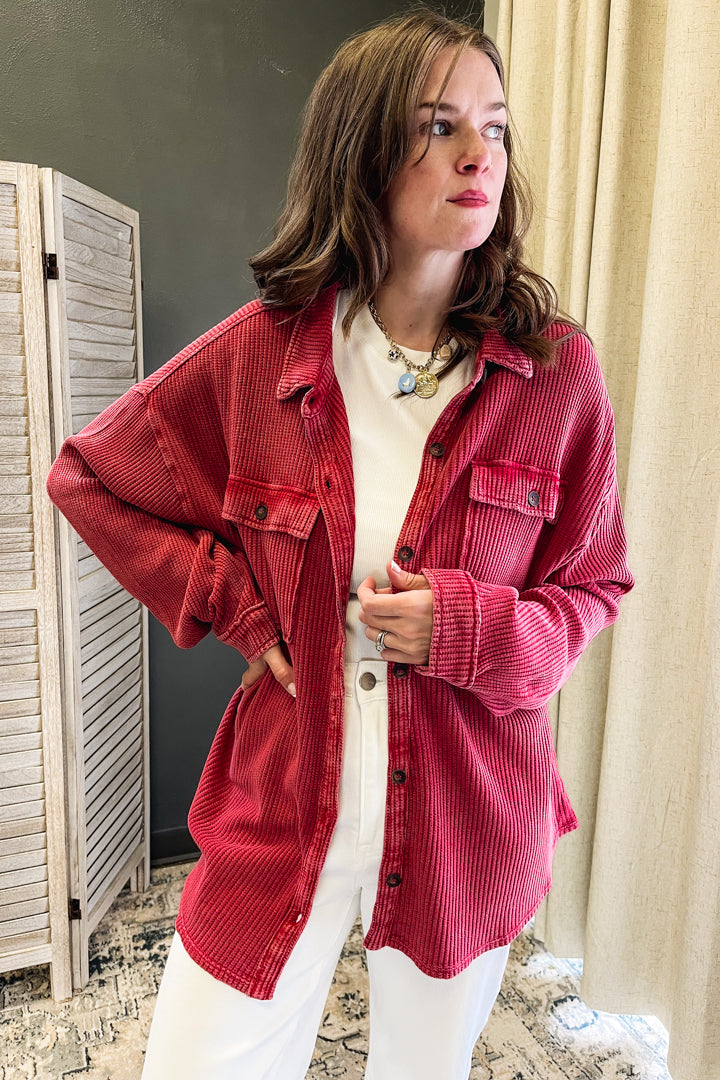 A woman wearing a red corduroy button-up shirt over a white top and white pants, standing in an indoor setting with a curtain and wooden screen visible.