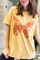 A woman wearing a mustard yellow t-shirt with a large butterfly graphic filled with orange, red, and white floral patterns, set against a softly blurred indoor background.