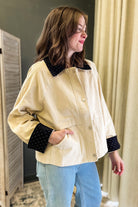 A woman wearing a cream-colored jacket with black polka-dot contrast trim on the collar and cuffs, paired with light blue jeans, standing indoors.
