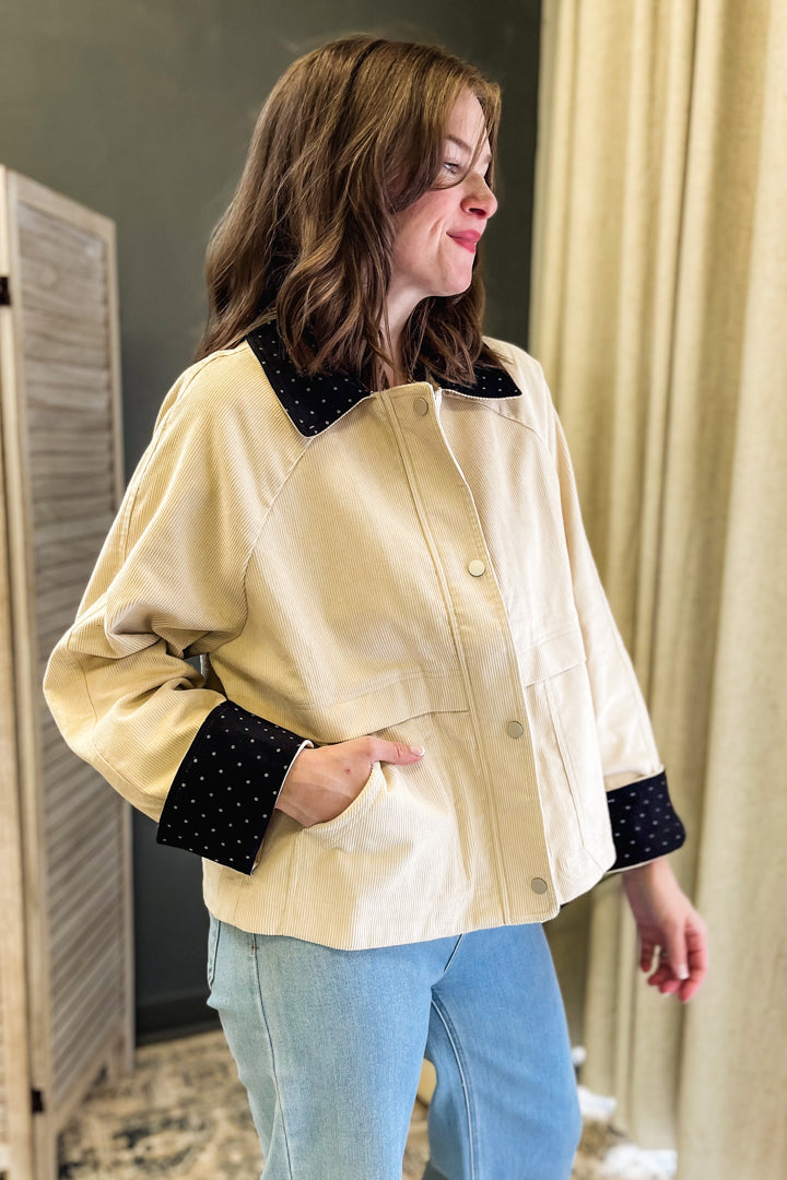 A woman wearing a cream-colored jacket with black polka-dot contrast trim on the collar and cuffs, paired with light blue jeans, standing indoors.