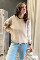A woman wearing a beige knit sweater with dark brown trim and light blue jeans, standing in an indoor setting with a folding screen and curtains in the background.