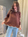 A woman wearing a brown knit sweater with cream ribbed trim at the crew neck, cuffs, and scalloped hem, paired with light blue jeans in an indoor setting.