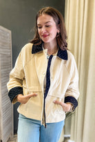 A woman wearing a cream-colored utility jacket with dark navy polka-dot collar and cuffs, paired with light blue jeans, standing indoors.
