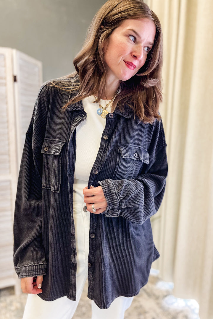 A woman wearing an oversized black corduroy button-up shirt jacket over a white top and white pants, standing indoors against a neutral beige curtain background.