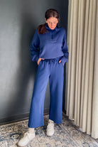 A woman wearing a matching blue lounge set consisting of a half-zip long-sleeve top and wide-leg drawstring pants, paired with white sneakers, standing in an indoor setting with a dark gray wall and beige curtains.