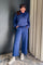 A woman wearing a matching blue lounge set consisting of a half-zip long-sleeve top and wide-leg drawstring pants, paired with white sneakers, standing in an indoor setting with a dark gray wall and beige curtains.