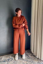 A woman wearing a rust-orange sweatshirt and matching wide-leg pants, standing in a room with a dark gray wall, beige curtains, and a patterned rug.