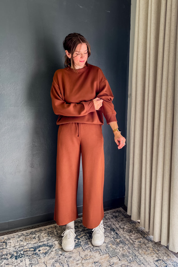 A woman wearing a rust-orange sweatshirt and matching wide-leg pants, standing in a room with a dark gray wall, beige curtains, and a patterned rug.