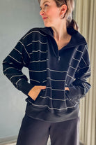 A woman wearing a black half-zip sweatshirt with thin white horizontal stripes and a stand-up collar, paired with black leggings, standing indoors against a gray wall.