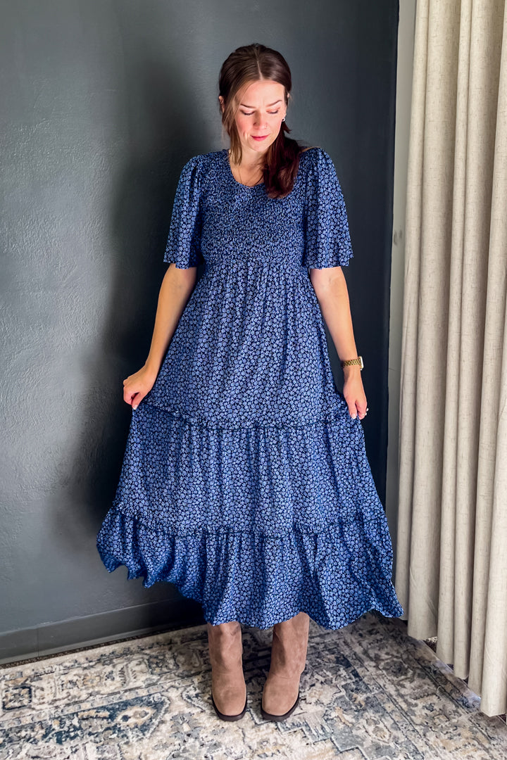 A woman wearing a blue tiered maxi dress with a small white floral print, standing in front of a dark gray wall and beige curtains.