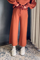 A person wearing rust-orange wide-leg sweatpants with a drawstring waist, paired with a matching sweatshirt, white socks, and white sneakers, standing on a patterned rug.