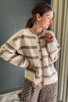 A woman wearing a cream-colored knit cardigan with brown and muted blue-gray horizontal stripes, paired with brown polka-dot pants, standing in an indoor setting.