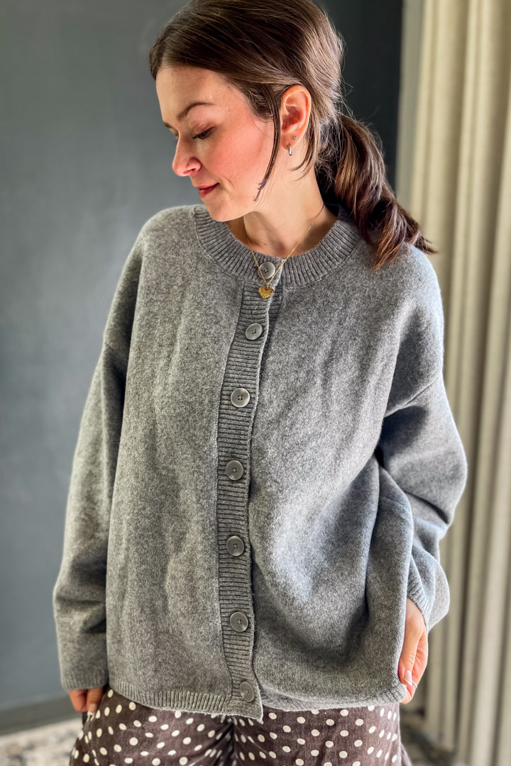 A woman wearing a gray knit cardigan sweater with a button front, shown in an indoor setting.