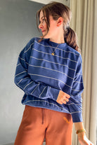 A woman wearing a blue long-sleeve sweatshirt with thin white horizontal stripes and rust-brown drawstring sweatpants, standing indoors against a gray wall and beige curtain.