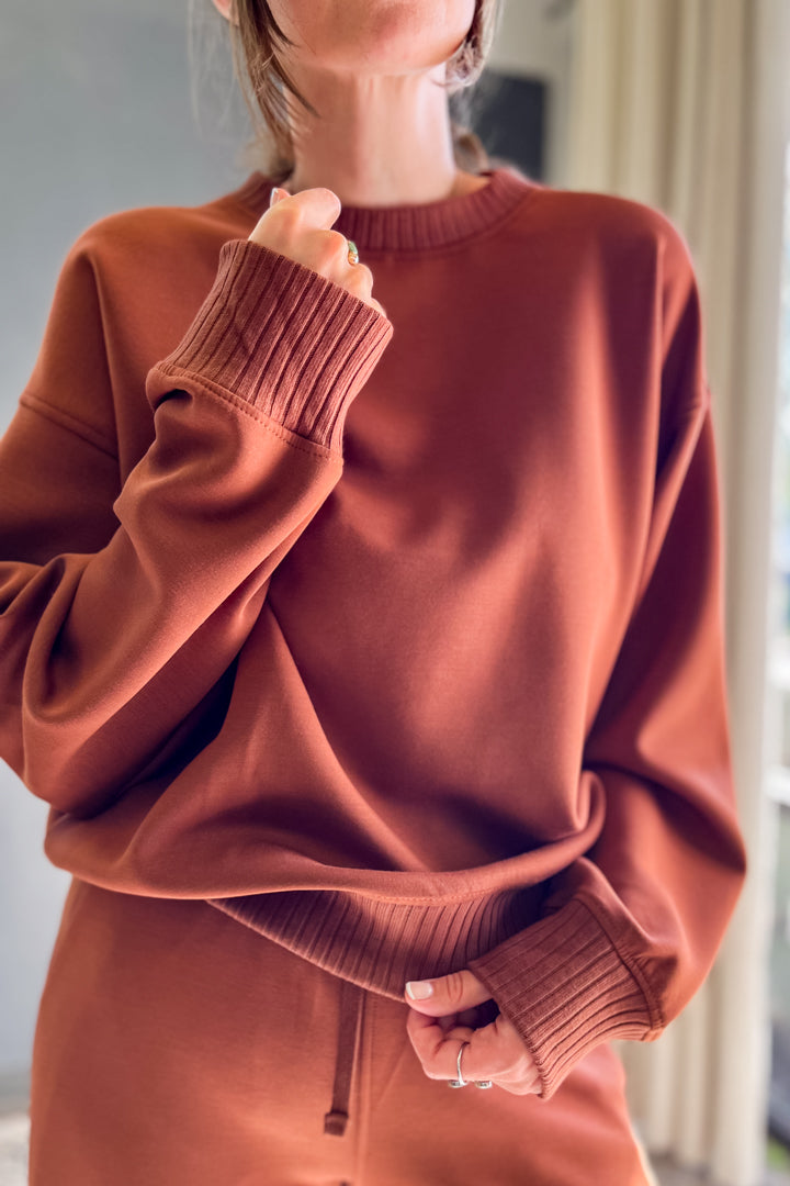 A person wearing an oversized ribbed crewneck sweatshirt in a warm rust or terracotta color, paired with matching ribbed bottoms. The photo is cropped from the lower face to the hips, set in a softly lit indoor environment.